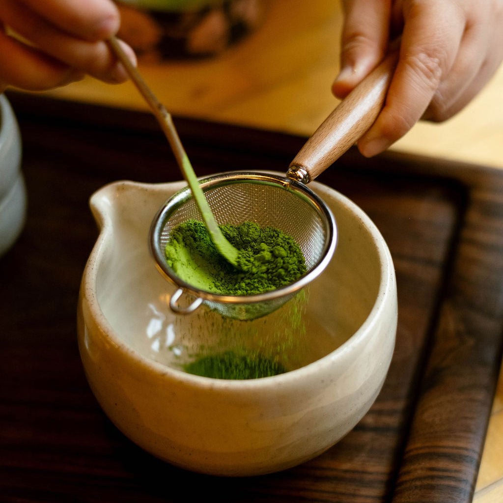 Person sifting green tea into a ceramic teapot using a wooden spoon.
